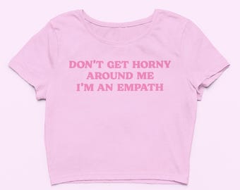 May include: A light pink crop top with the text "DON'T GET HORNY AROUND ME I'M AN EMPATH" in a darker pink. The shirt has a crew neck and short sleeves. The text is centered on the front of the shirt.