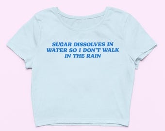 May include: A light blue crop top with the text "SUGAR DISSOLVES IN WATER SO I DON'T WALK IN THE RAIN" in blue. The shirt has short sleeves and a round neckline. The background is a light pink.