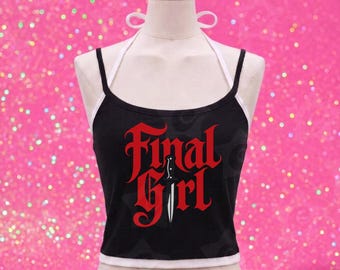 May include: Black tank top with the words "Final Girl" in red gothic lettering and a knife graphic. The top has a white trim and straps, displayed on a mannequin against a pink glitter background.