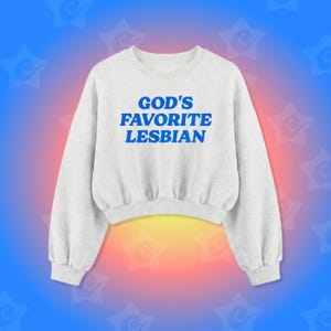 May include: A pale grey sweatshirt featuring the blue text "GOD'S FAVORITE LESBIAN". The cropped top has long sleeves. The background is a gradient of blue, red, and yellow with star shapes.