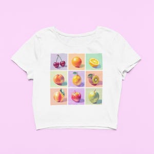 May include: White crop top featuring a colorful grid of painted fruits: cherries, orange, lemon, peach, lemon, kiwi, peach, apple, and green apple. The design is set against a pastel background, creating a vibrant and artistic aesthetic.