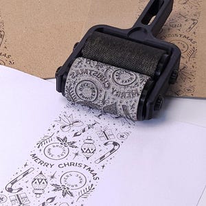 Può includere: Un timbro a rullo nero con un design a tema natalizio viene mostrato, che rotola su carta bianca. Il timbro crea un motivo ripetuto di icone festive e le parole "Merry Christmas". Una busta marrone è sullo sfondo.