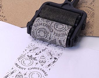 Könnte beinhalten: Ein schwarzer Walzenstempel mit weihnachtlichem Design wird gezeigt, der über weißes Papier rollt. Der Stempel erzeugt ein sich wiederholendes Muster aus Feiertags-Symbolen und den Worten "Merry Christmas". Ein brauner Umschlag ist im Hintergrund.