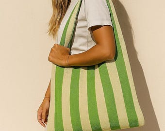 May include: A green and cream striped tote bag with long straps. The bag is made of a woven material and is being held by a person. The bag is a light green color with cream-colored stripes. The bag is a good size for carrying items.