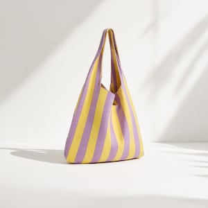 Könnte beinhalten: Eine weiche Stricktasche im Hobo-Stil mit vertikalen Streifen in abwechselndem hellem Gelb und zartem Lavendel. Die Tasche hat eine dreieckige Form mit einem integrierten Griff an der Oberseite und besteht aus einem flexiblen, strukturierten Stoff.