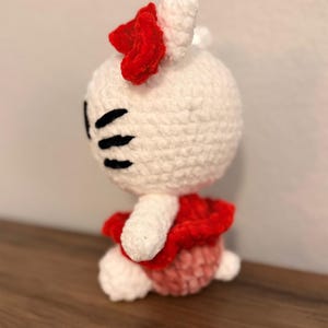 May include: A handmade crochet doll in the shape of a cartoon cat. The doll is primarily white with black eyes and whiskers. It wears a red bow and a red and pink dress. The doll is sitting on a wooden surface.