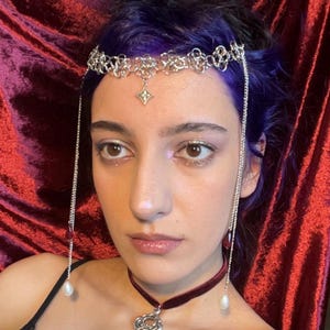 May include: A silver chainmail headpiece featuring a central star-shaped charm and two long side chains ending in red teardrop beads and white pearls. A dark red velvet choker necklace with a silver key pendant is worn around the neck.