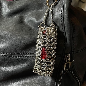 Może przedstawiać: Srebrny urok z kolczugi z czerwoną kryształową zawieszką, wiszący na czarnej skórzanej torbie. Urok jest prostokątny, a kryształ w kształcie łzy. Torba ma zamek błyskawiczny i złoty pierścień.