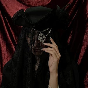 May include: A black wide-brimmed hat with a feathered accent and a long black lace veil covering the face. A silver-toned chain necklace with a decorative filigree pendant rests over the lace veil, and a silver-toned chain bracelet is worn on the wrist.