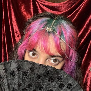 May include: A silver-toned head chain featuring delicate metal links and a central decorative filigree element, worn across the forehead. The accessory is paired with a black folding hand fan decorated with black floral embroidery and sequins.