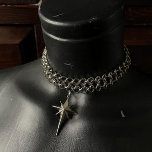 May include: A silver-toned chainmail choker necklace featuring a polished metal star-shaped pendant with eight points. The choker is constructed from interlocking circular metal rings, creating a woven pattern that sits flush against the neck.