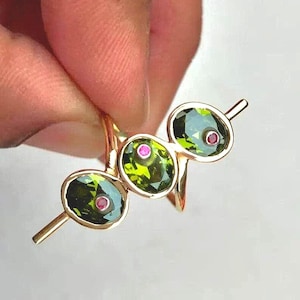 May include: A gold ring with three oval green gemstones, each with a small pink stone at the centre. The ring is held up, showing its design and colour against a white background. The gemstones are set in a gold band.