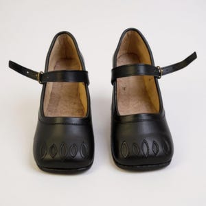 May include: A pair of black leather Mary Jane style shoes. Each shoe has a strap with a buckle closure and decorative stitching on the toe. The shoes are set against a white background.