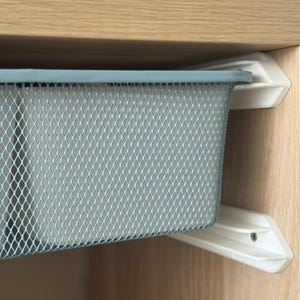 May include: A light blue rectangular wire mesh storage basket mounted on white plastic sliding rails. The basket features a diamond-patterned metal grid design and is positioned horizontally within a light-colored wooden cabinet structure.