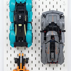 May include: Three toy cars constructed from interlocking plastic bricks are mounted on a white pegboard. One car is bright turquoise, one is dark gray with orange accents, and the third is orange with blue flame graphics and black wheels.