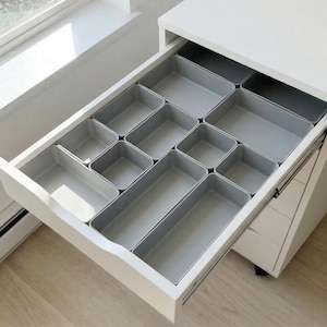 May include: A white desk drawer with a set of gray storage organizers. The drawer is partially open, revealing the compartments. A small clock and potted plant sit on the windowsill in the background.