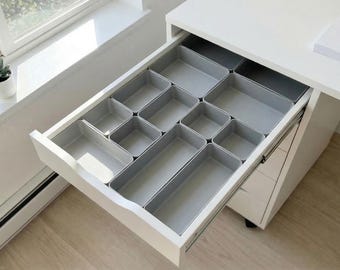 May include: A white desk drawer with a set of gray storage organizers. The drawer is partially open, revealing the compartments. A small clock and potted plant sit on the windowsill in the background.
