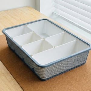 May include: A rectangular desk organizer tray featuring a dark gray metal mesh outer frame and six removable white plastic interior compartments of varying sizes, designed for sorting small office supplies and stationery items efficiently.