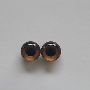 Könnte beinhalten: Zwei runde Puppenaugen aus Glas mit einer dunkelbraunen Iris und einem hellbraunen Außenring. Die Augen haben eine glänzende Oberfläche und sind vor einem schlichten weißen Hintergrund platziert. Diese Bastelmaterialien sind ideal für die Puppenherstellung.
