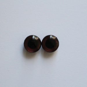 May include: A pair of brown doll eyes with a dark pupil and a shiny surface. The eyes are circular and likely made of glass or a similar material. They are positioned on a plain white backdrop.