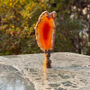 May include: A lamp finial featuring a polished, orange agate slice with a vibrant red center. The agate is mounted on a brass base with a detailed, textured design. The finial is set against a blurred green background.