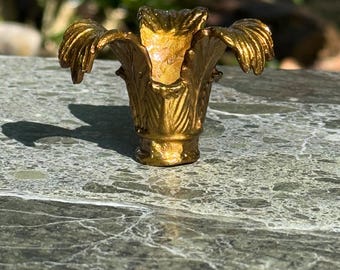 May include: A gold-colored, ornate decorative object, possibly a bottle stopper or small vase. The object has a floral design with detailed petal-like structures and a flared base. The surface has a metallic sheen, reflecting light.