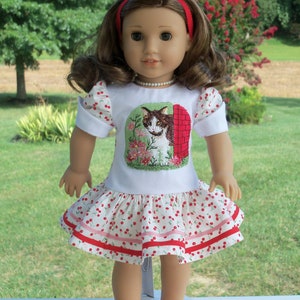 May include: A doll wearing a white dress with red and white cherry print and a red headband. The dress has a white embroidered image of a cat in front of a brick building. The doll is wearing red shoes.