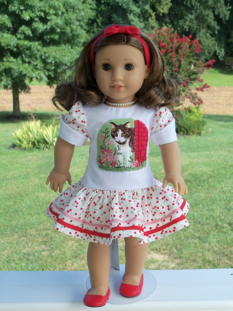 May include: A doll wearing a white dress with red and white cherry print and a red headband. The dress has a white embroidered image of a cat in front of a brick building. The doll is wearing red shoes.