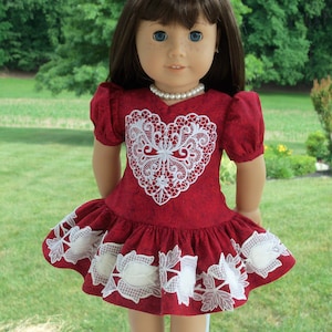 May include: A red dress with white lace detailing in the shape of a heart. The dress has a ruffled hem and is worn by a doll with brown hair and blue eyes.