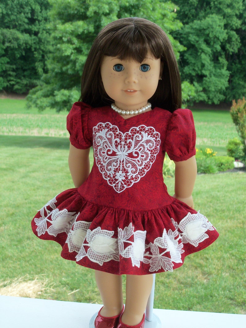 May include: A red dress with white lace detailing in the shape of a heart. The dress has a ruffled hem and is worn by a doll with brown hair and blue eyes.