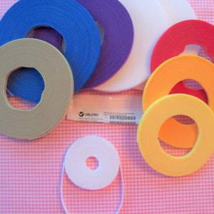 May include: Assortment of colorful hook and loop tape rolls in various sizes. The rolls are arranged in a circle on a pink and white gingham background. The tape is labeled "VELCRO" and includes a product code.