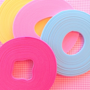 May include: Four spools of colourful ribbon in shades of yellow, pink, and blue are arranged on a pink and white checkered background. The ribbon is tightly wound, with a hole in the centre of each spool. The image is brightly lit and has a cheerful aesthetic.