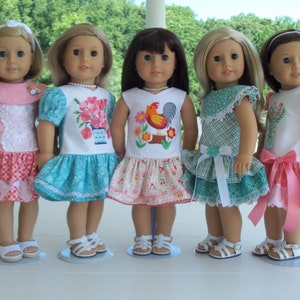 May include: Five dolls wearing different dresses with embroidered designs. The dolls are all wearing white sandals and are standing on a white surface.