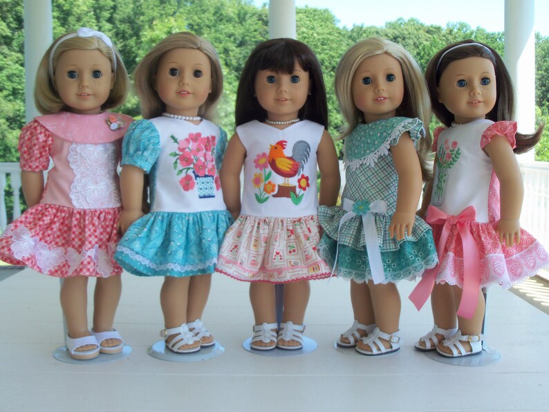 May include: Five dolls wearing different dresses with embroidered designs. The dolls are all wearing white sandals and are standing on a white surface.
