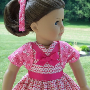 May include: A pink and white dress with a pink bow and a matching pink cardigan. The dress has a diamond pattern and the cardigan has a floral pattern.