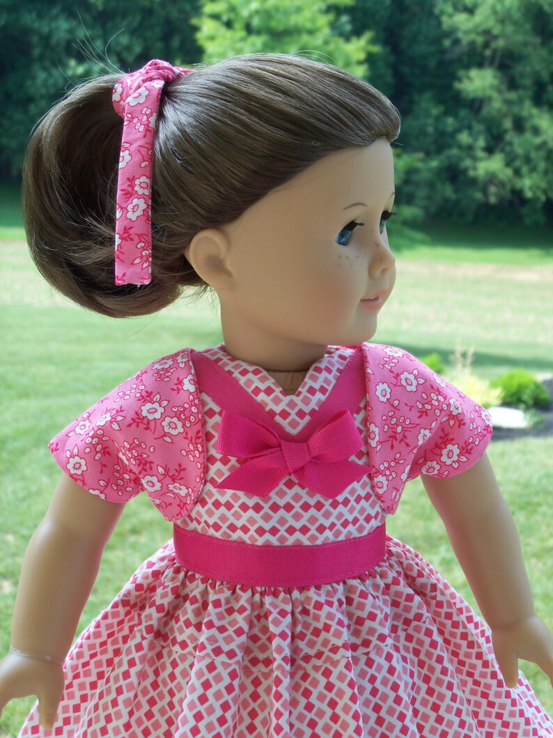 May include: A pink and white dress with a pink bow and a matching pink cardigan. The dress has a diamond pattern and the cardigan has a floral pattern.