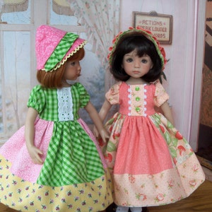 May include: Two dolls wearing pink and green dresses with floral patterns. The doll on the left is wearing a pink and white hat, and the doll on the right is wearing a pink hat with a white trim. Both dolls have long brown hair.