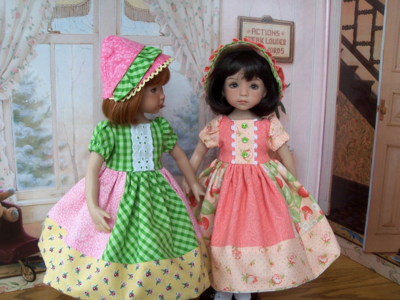 May include: Two dolls wearing pink and green dresses with floral patterns. The doll on the left is wearing a pink and white hat, and the doll on the right is wearing a pink hat with a white trim. Both dolls have long brown hair.