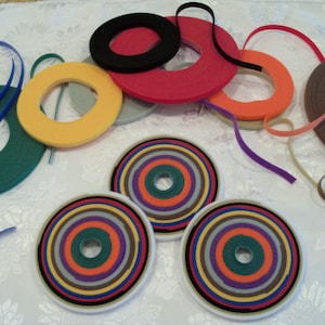 May include: Assortment of colorful fabric ribbon rolls in various widths and colors, including red, orange, yellow, green, blue, brown, and black. The rolls are arranged on a white surface.