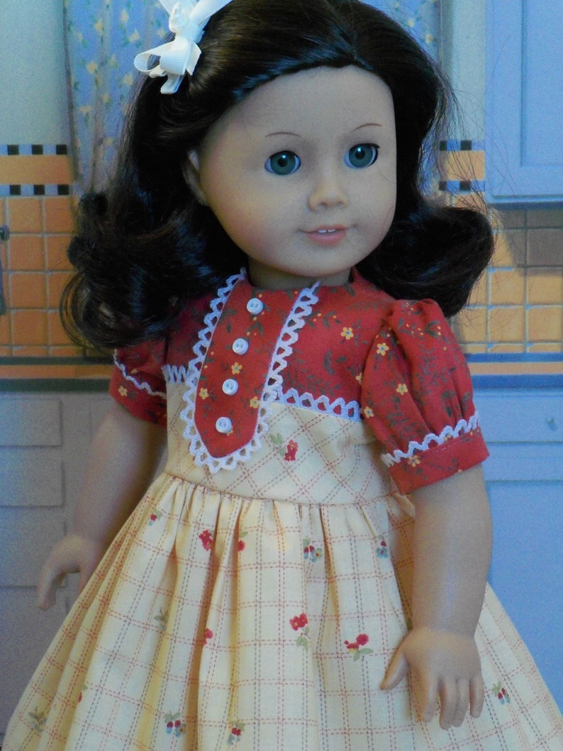 May include: A doll wearing a yellow and red dress with a white lace trim. The dress has a plaid pattern and small red flowers. The doll has long dark hair and blue eyes.