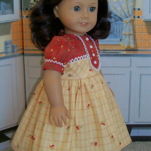 May include: A doll wearing a yellow and red plaid dress with a white lace trim and a red bow in her hair. The doll is standing on a white surface.