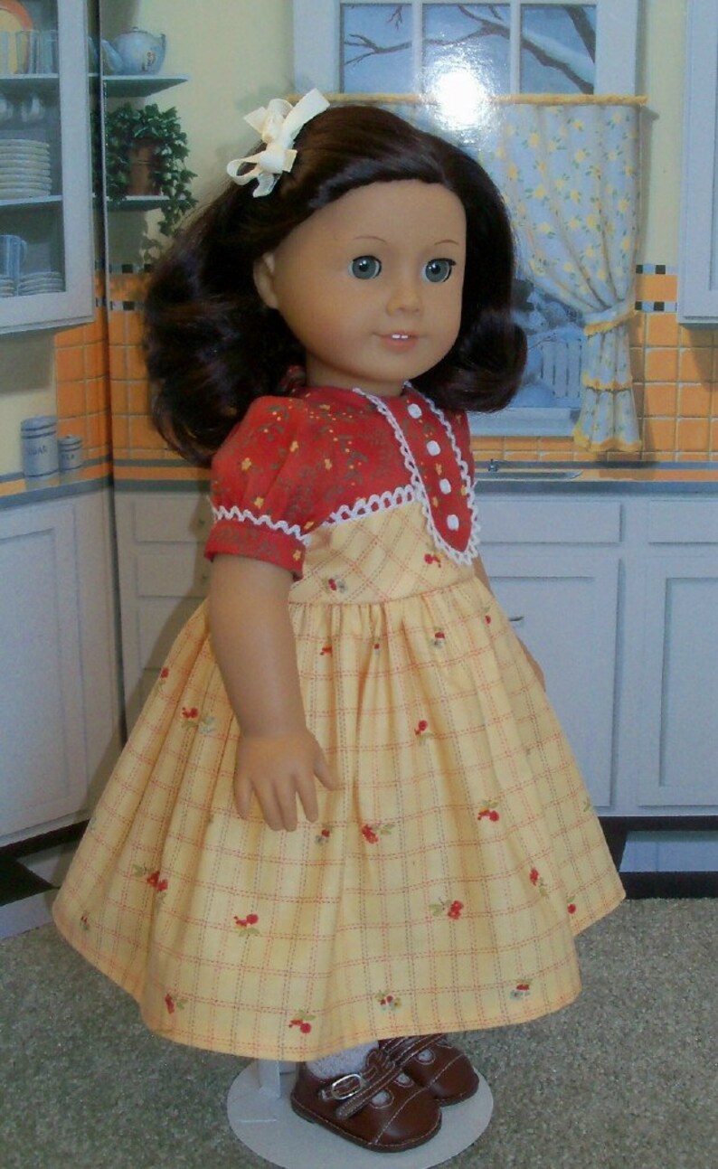 May include: A doll wearing a yellow and red plaid dress with a white lace trim and a red bow in her hair. The doll is standing on a white surface.