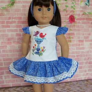 May include: A blue and white dress with a bird and birdbath embroidery design. The dress has a white lace trim and is worn by a doll with brown hair and blue eyes.