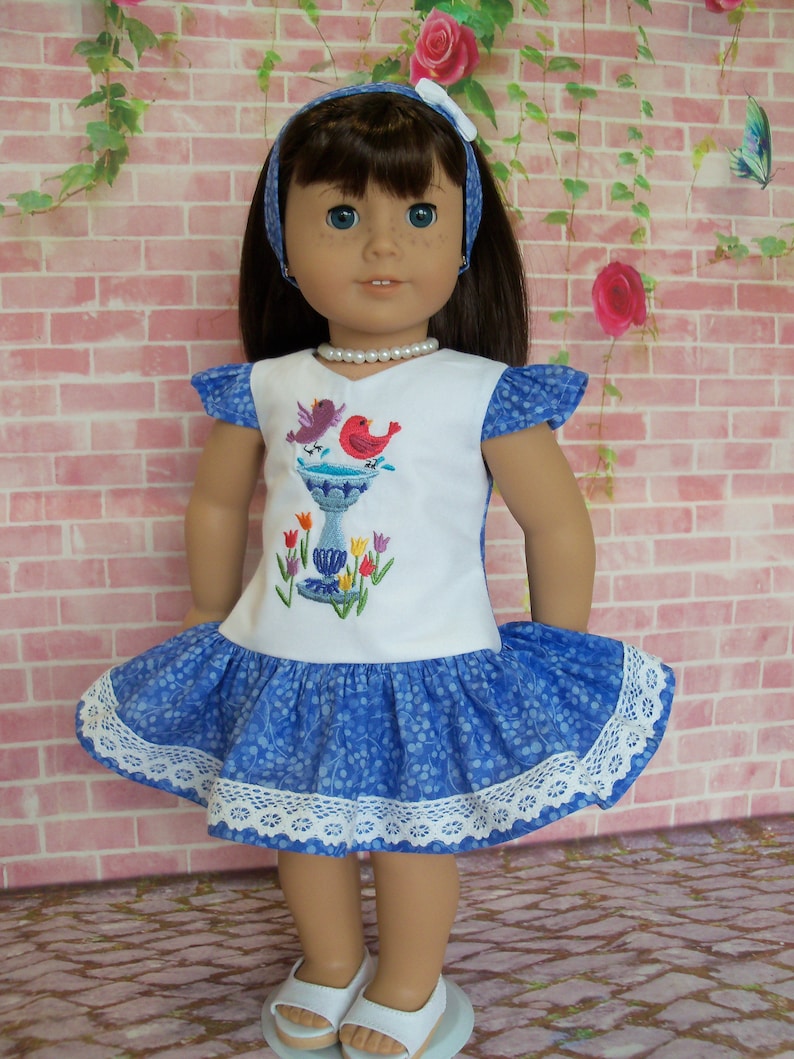 May include: A blue and white dress with a bird and birdbath embroidery design. The dress has a white lace trim and is worn by a doll with brown hair and blue eyes.