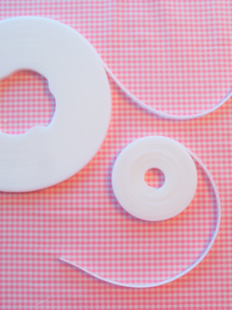 May include: Two spools of white elastic ribbon, one large and one small, are positioned on pink and white gingham fabric. The ribbon is unrolled from the spools, forming a thin white line.