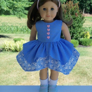 May include: A blue and white gingham dress with a white floral print hem. The dress has pink heart-shaped buttons down the front. The doll is wearing white socks and blue shoes.