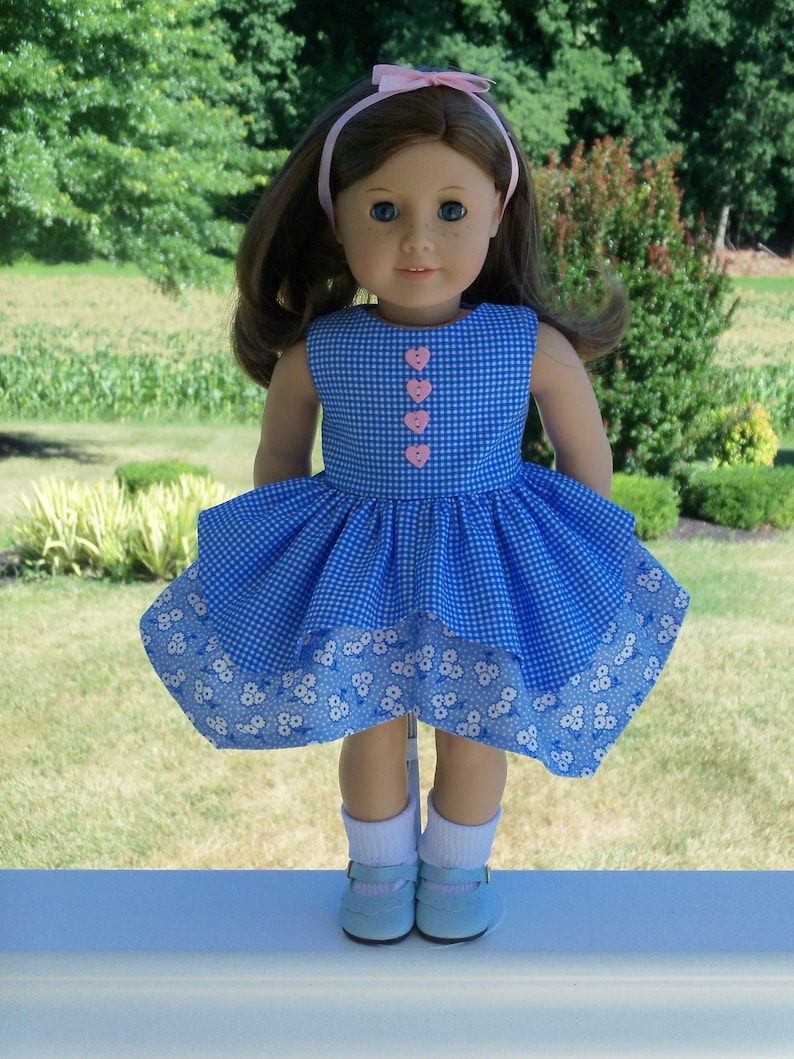 May include: A blue and white gingham dress with a white floral print hem. The dress has pink heart-shaped buttons down the front. The doll is wearing white socks and blue shoes.