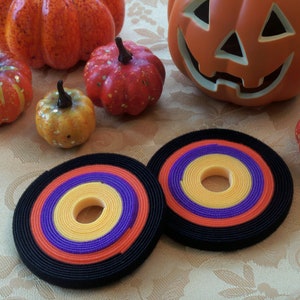 May include: Two black, orange, yellow, and purple striped cable ties with a hole in the center. The ties are on a white tablecloth with small pumpkins and a jack-o-lantern.