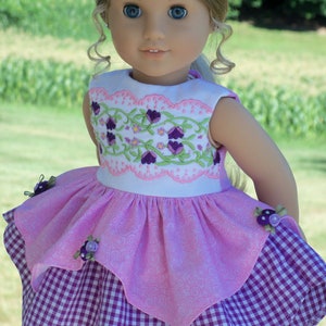 May include: A doll wearing a pink and purple dress with a floral embroidered bodice and a gingham skirt. The doll has blonde hair and blue eyes and is wearing a flower crown.