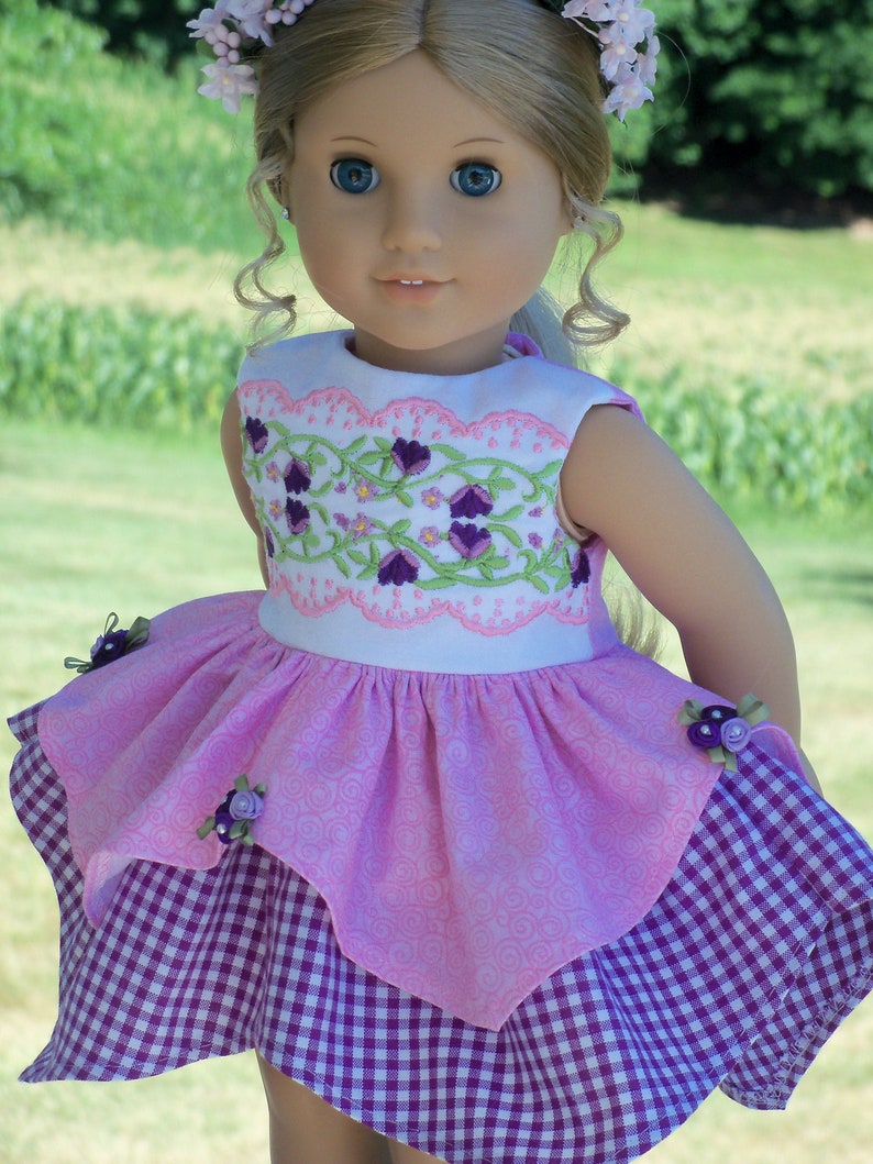 May include: A doll wearing a pink and purple dress with a floral embroidered bodice and a gingham skirt. The doll has blonde hair and blue eyes and is wearing a flower crown.